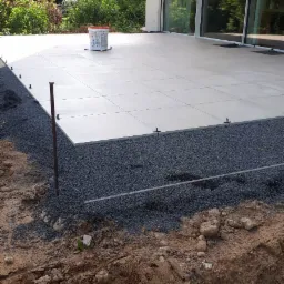 Montaż tarasu betonowego: płyty ułożone na warstwie żwiru z widocznymi elementami dystansowymi, w tle budynek z dużymi oknami i zieleń.