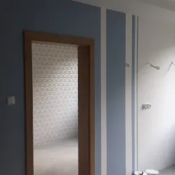 Wnętrze pokoju w trakcie wykańczania: ściana pomalowana na niebiesko z białymi pasami, obok otwarta futryna z widoczną tapetą w geometryczny wzór, biała ściana z wystającymi przewodami...