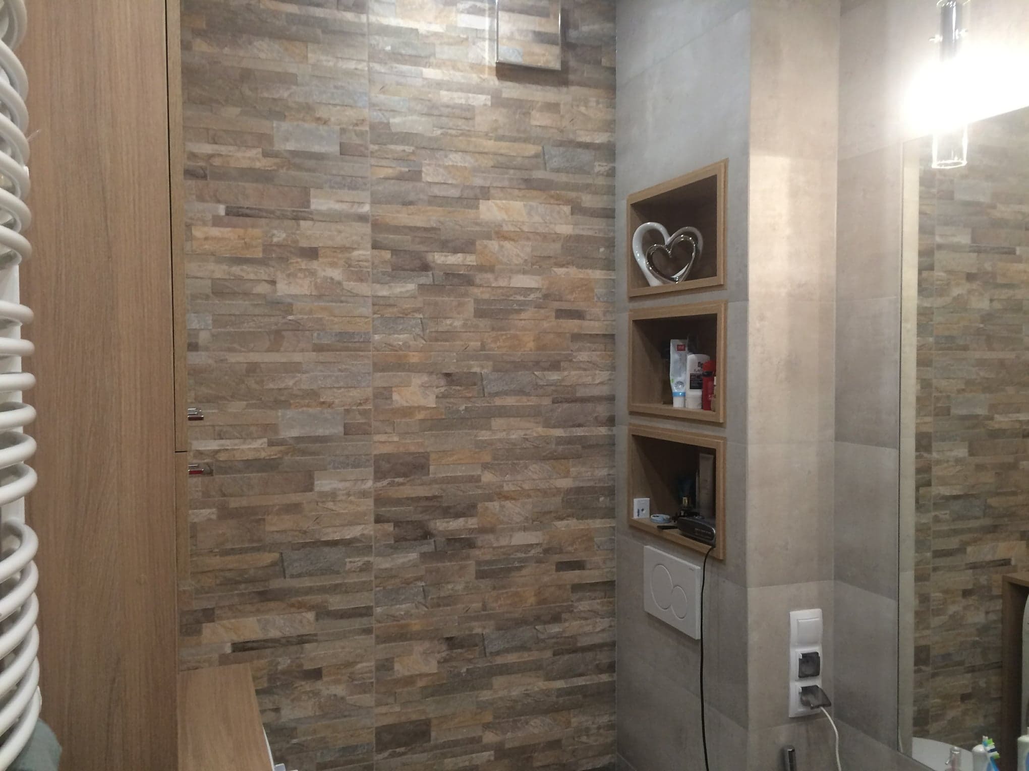Wykończona łazienka z kamienną ścianą, drewnianymi półkami z dekoracją w kształcie serca i lustrem, widoczne gniazdka elektryczne i przycisk spłuczki.