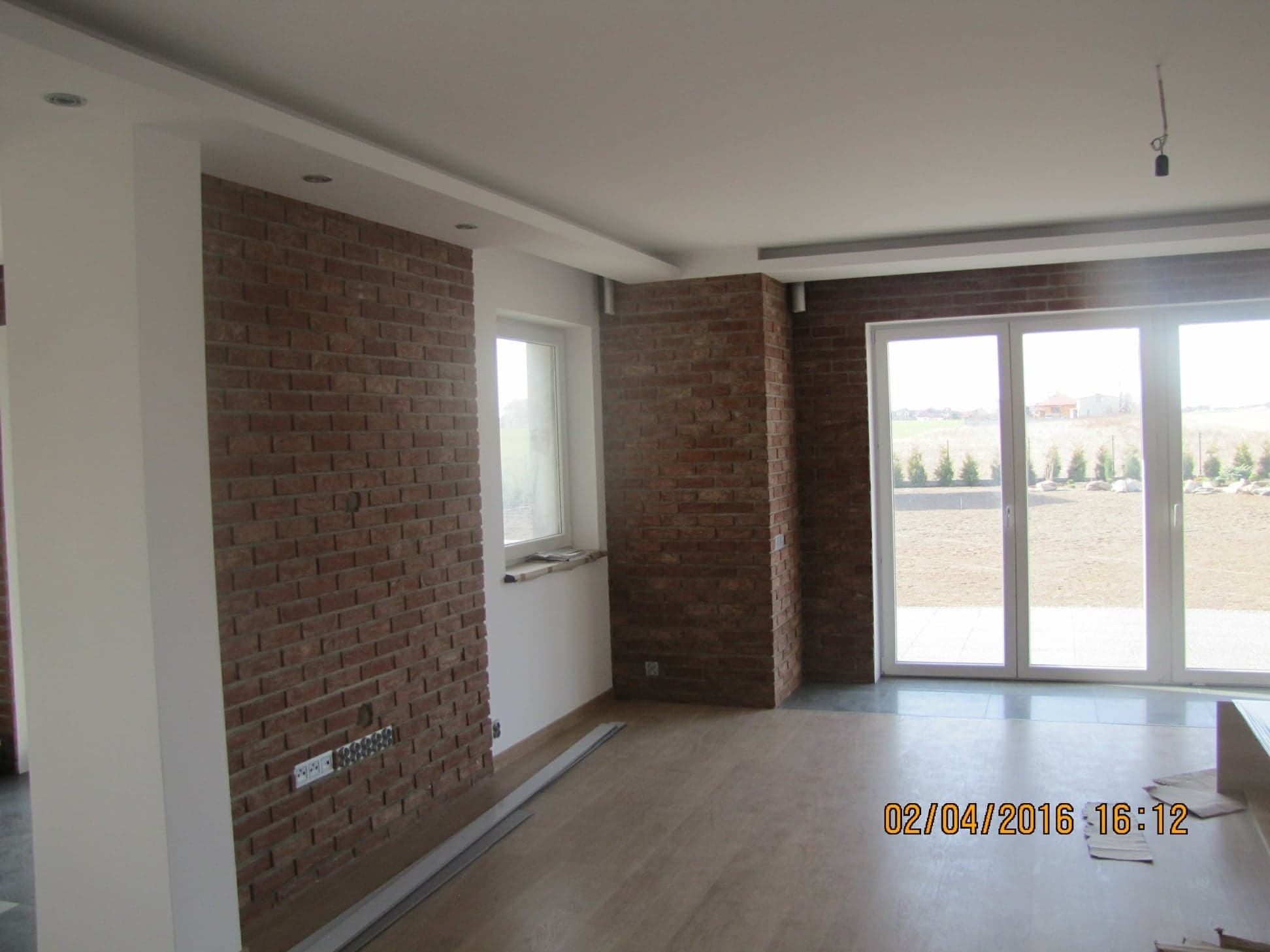 Salon z częściowo wykończonymi ścianami z czerwonej cegły, widoczne gniazdka elektryczne, duże okna z widokiem na ogród, jasna podłoga i listwy przypodłogowe.