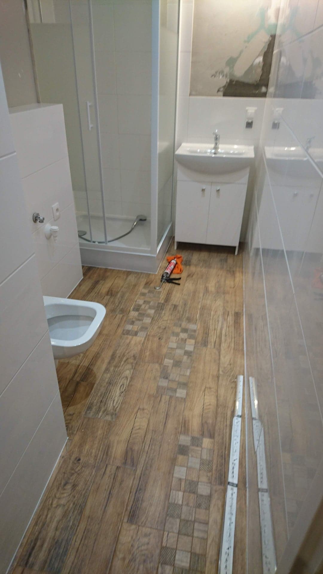 Wykończona łazienka z drewnopodobnymi płytkami podłogowymi z dekoracyjnymi wstawkami, białą armaturą, kabiną prysznicową i narzędziami do fugowania w rogu.