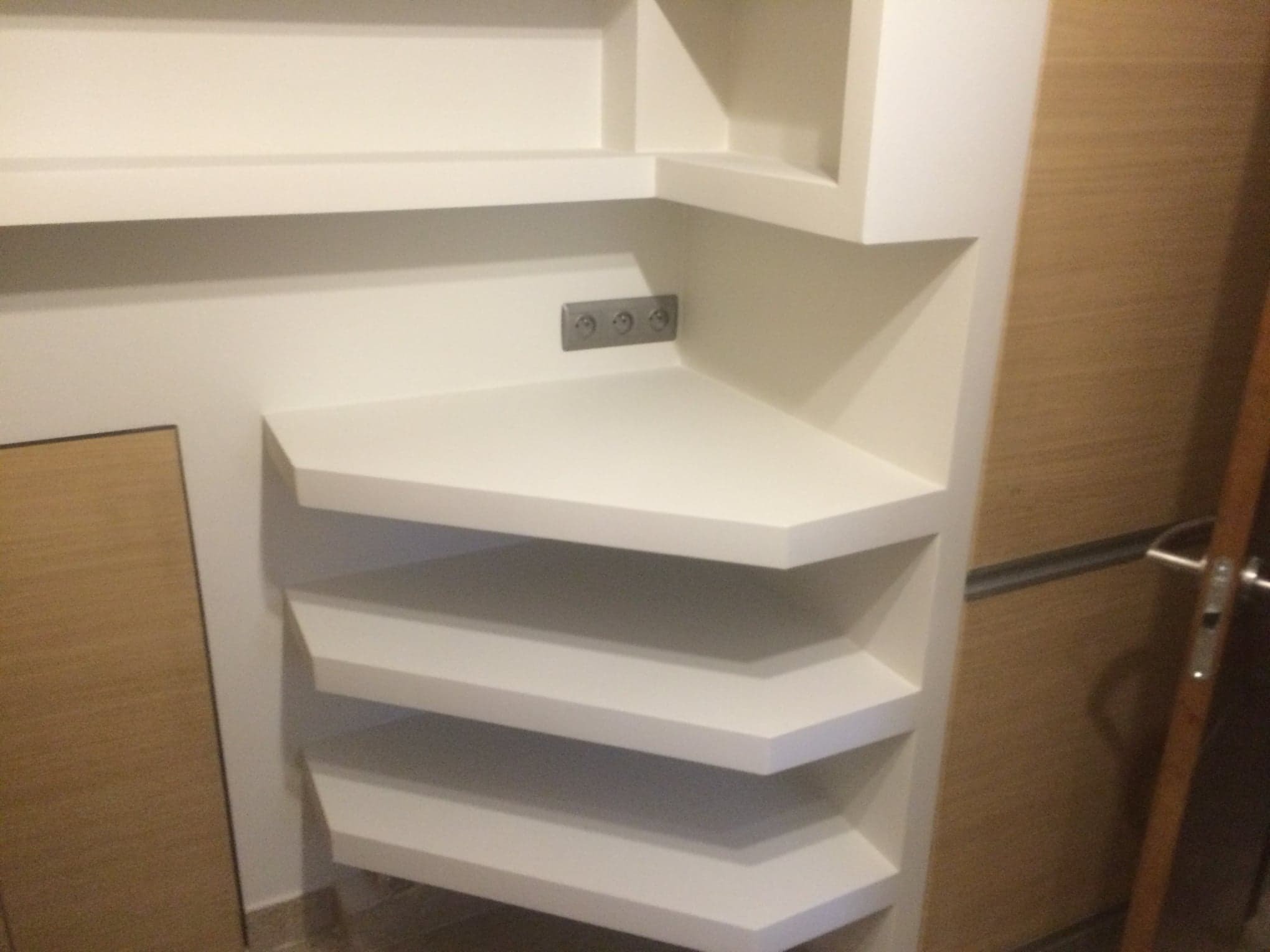 Biała, narożna zabudowa z półkami, wbudowane gniazdka elektryczne, widok na fragment drzwi.