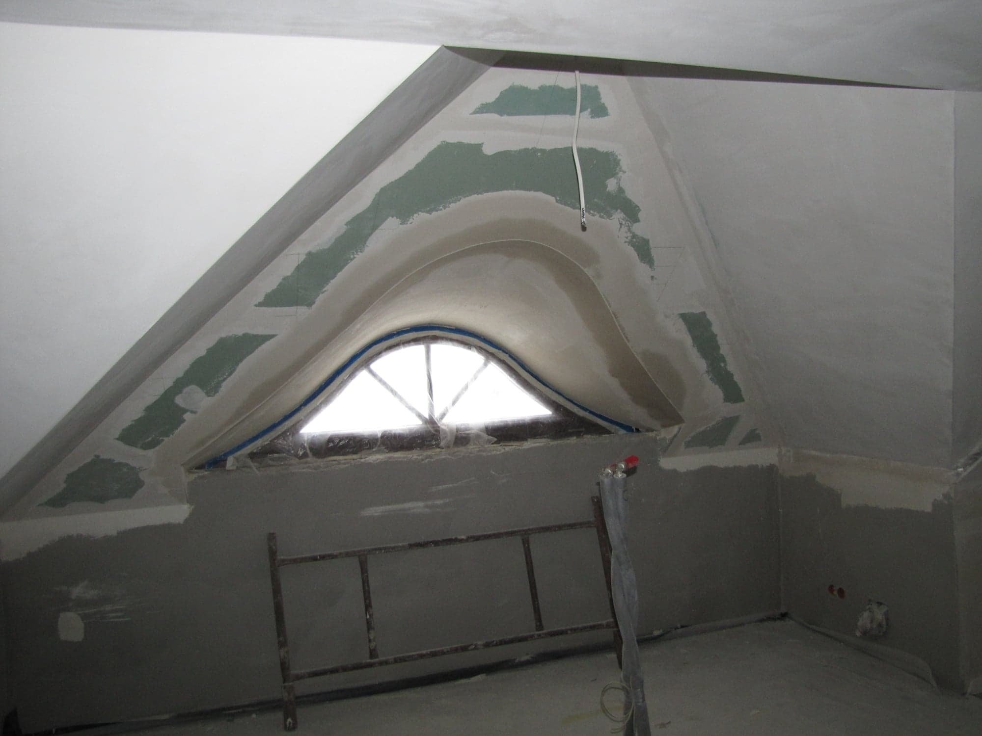 Poddasze w trakcie remontu, widoczne szpachlowane ściany, okno w kształcie łuku oraz drabina malarska.