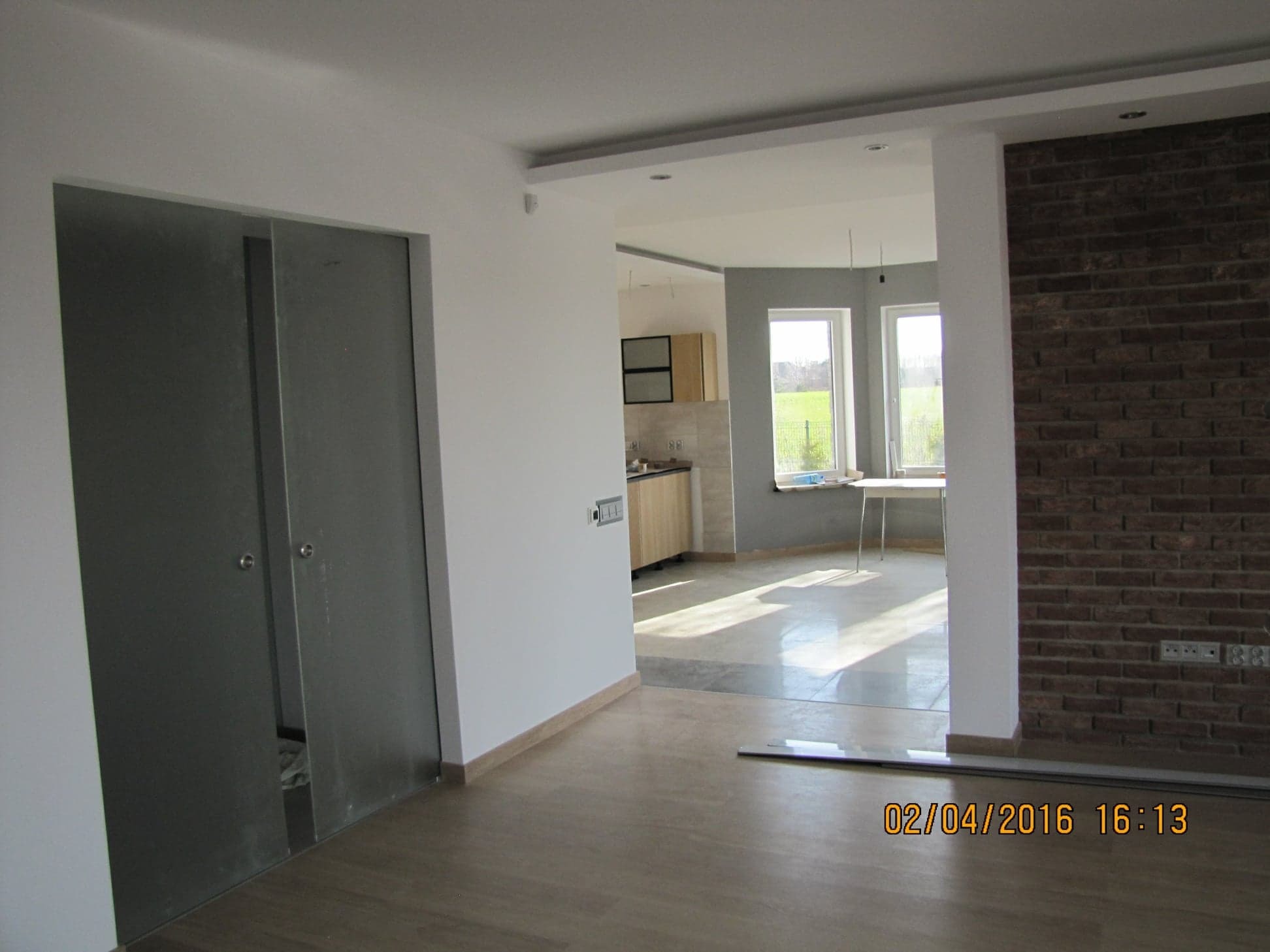 Wnętrze mieszkania po remoncie: widok na kuchnię z salonu, przesuwne drzwi częściowo otwarte, ściana z cegły dekoracyjnej, drewniana podłoga.