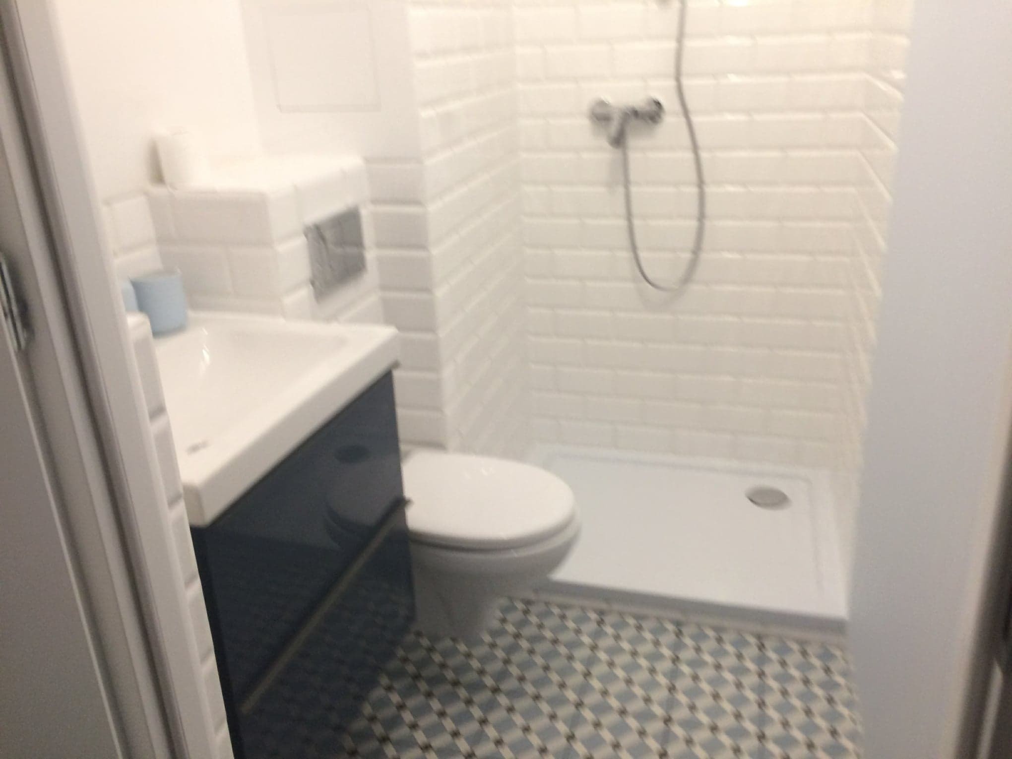 Wyremontowana łazienka z kabiną prysznicową, umywalką z szafką w kolorze granatowym i toaletą, wykończona białą cegiełką i wzorzystą podłogą.