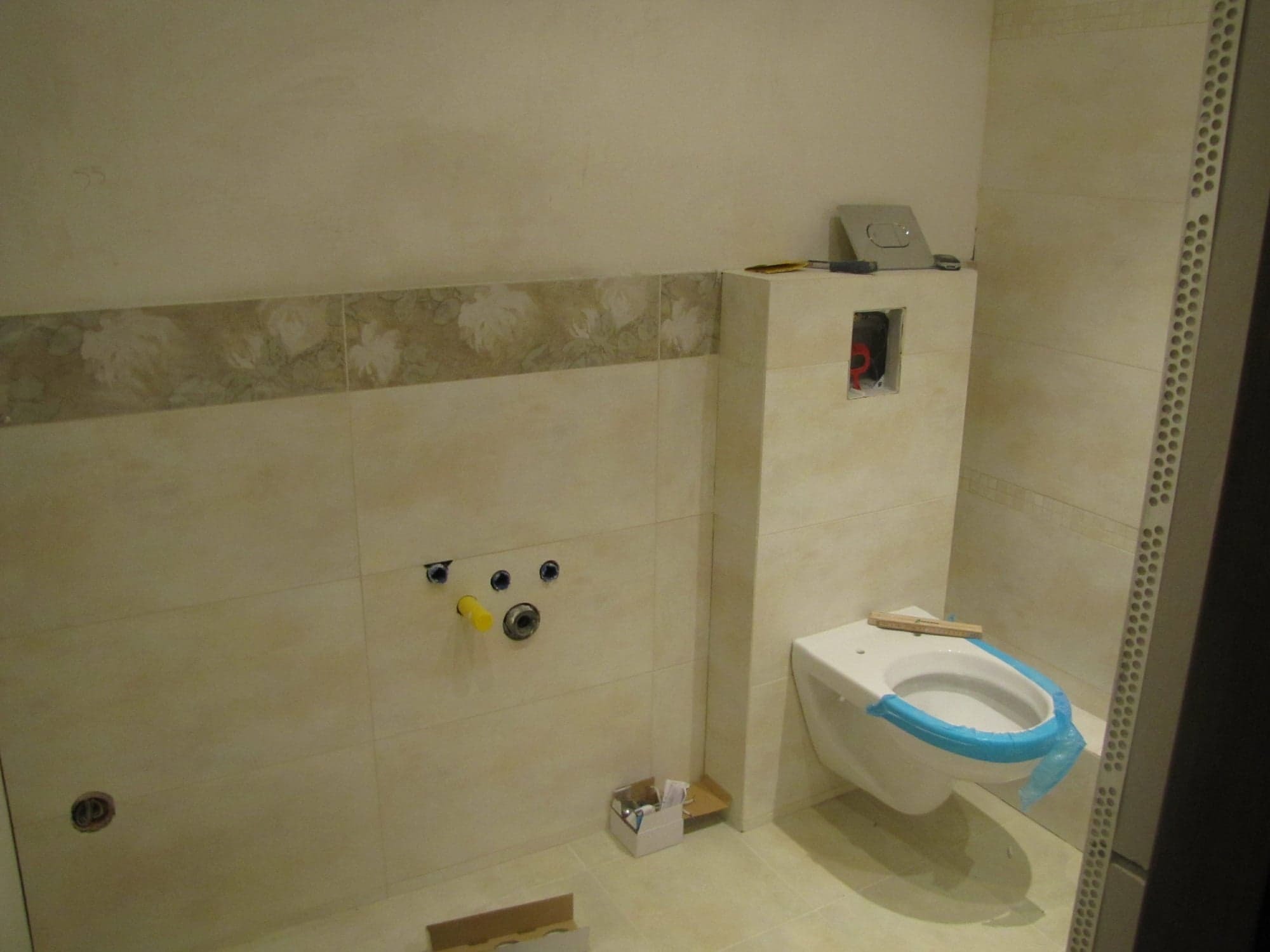 Wnętrze łazienki w trakcie wykańczania: podwieszana toaleta z niebieskim zabezpieczeniem, częściowo ułożone płytki z dekoracyjnym pasem, widoczne przyłącza wodno-kanalizacyjne i puszka pod przycisk...