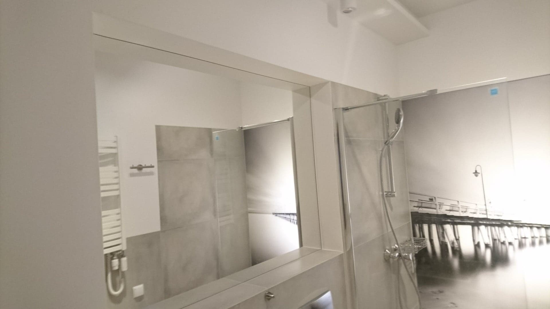 Nowoczesna łazienka z dużym lustrem, grzejnikiem drabinkowym i kabiną prysznicową z deszczownicą, na ścianie fototapeta z motywem molo.