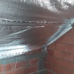 Poddasze w trakcie ocieplania, widoczna folia aluminiowa termoizolacyjna na skośnym dachu i ceglana ściana z instalacją elektryczną.
