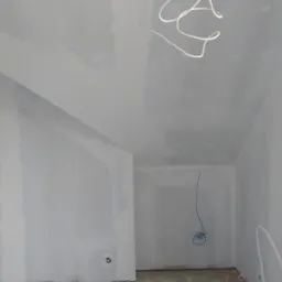 Wnętrze pokoju w trakcie prac wykończeniowych, ściany pokryte gładzią gipsową, widoczne przewody elektryczne zwisające z sufitu i wystające ze ściany, przygotowane do dalszych prac instalacyjnych.