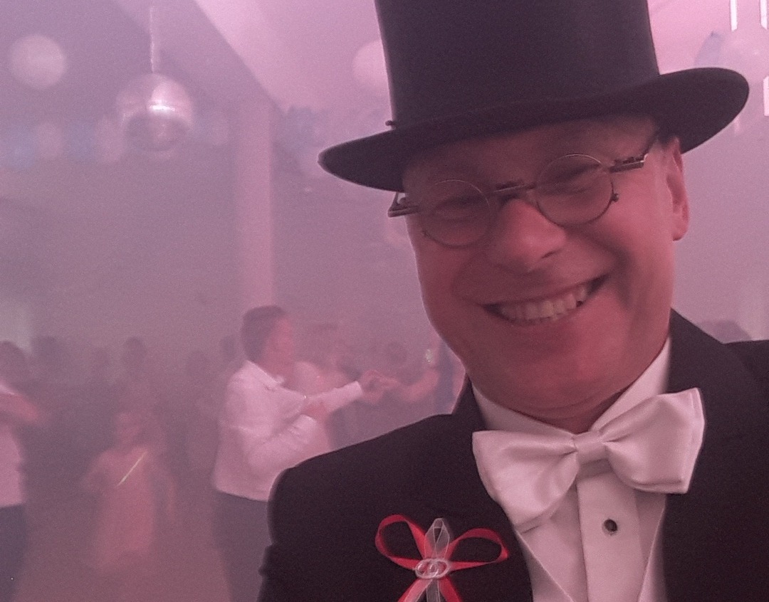 Uśmiechnięty mężczyzna w cylindrze i muszce, z przypinką z obrączkami, na tle sali tanecznej w Ciechanowie, oświetlonej dyskotekową kulą i mgłą.