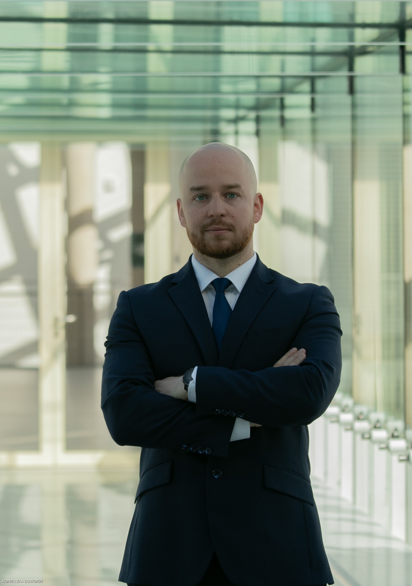 Portret mężczyzny w garniturze z założonymi rękami na tle przeszklonego korytarza. Profesjonalny wizerunek prawnika z brodą i zegarkiem.