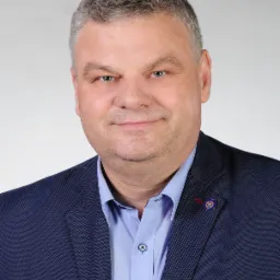 Portret uśmiechniętego mężczyzny w niebieskiej koszuli i marynarce z przypinką na klapie, na jasnym tle.