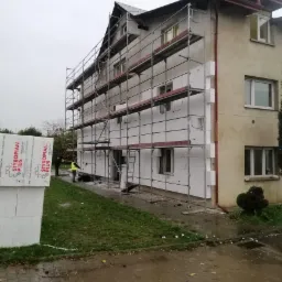 Trzy kondygnacyjny budynek w trakcie prac izolacyjnych, z widocznym rusztowaniem i częściowo zamontowanymi płytami styropianowymi, pracownik w ż&oacute;łtej kamizelce przycina płyty na parterze.