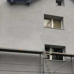 Szara elewacja budynku jednorodzinnego w trakcie prac wykończeniowych, widoczne rusztowanie z drabinką i fragment logo firmy na elewacji.