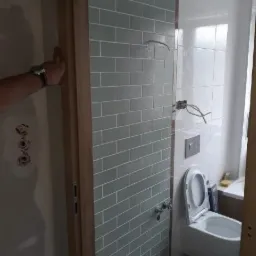 Wnętrze łazienki w trakcie prac wykończeniowych, widoczne szare, prostokątne płytki ułożone na ścianie, biała toaleta podwieszana, niezamaskowane przewody elektryczne oraz fragment dłoni...