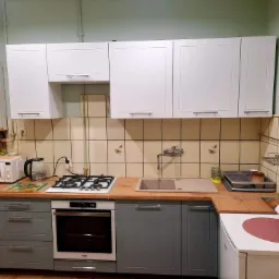 Wyposażenie kuchni po remoncie: szare szafki dolne z drewnianym blatem, białe szafki wiszące, kuchenka gazowa, piekarnik, zlew, bojler gazowy na ścianie. Na półce szklanej słoiki z przetworami.