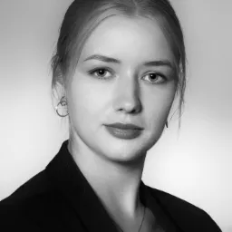 Czarno-białe portretowe zdjęcie młodej kobiety w ciemnej marynarce, z delikatną biżuterią i kolczykami, na jasnym tle, z subtelnym uśmiechem.