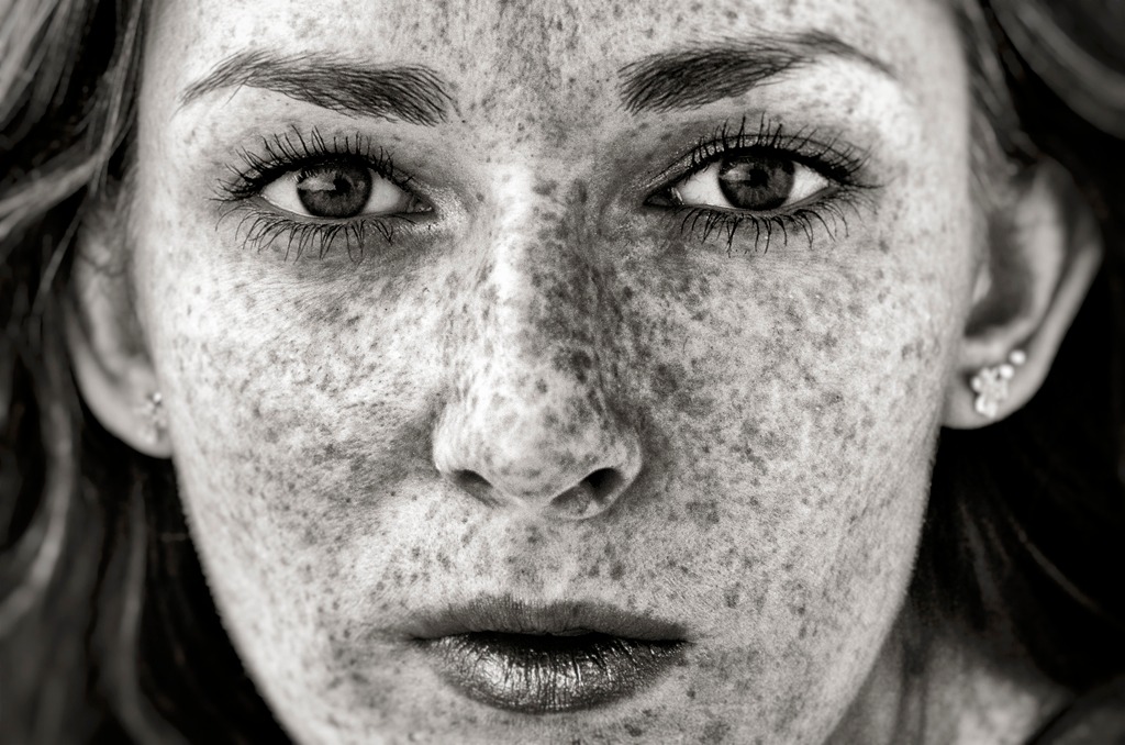 Czarno-biały portret kobiety z wyraźnymi piegami, intensywnym spojrzeniem i błyszczącymi ustami, uchwycony w bliskim ujęciu.