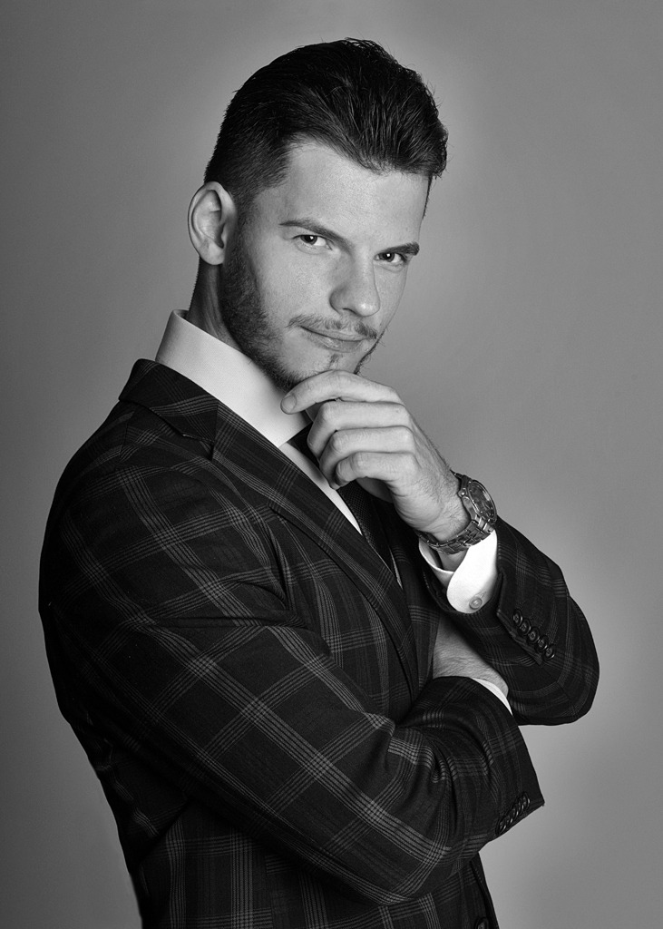 Czarno-białe portretowe ujęcie mężczyzny w garniturze w kratę, z ręką pod brodą i zegarkiem na nadgarstku, patrzącego z lekkim uśmiechem.