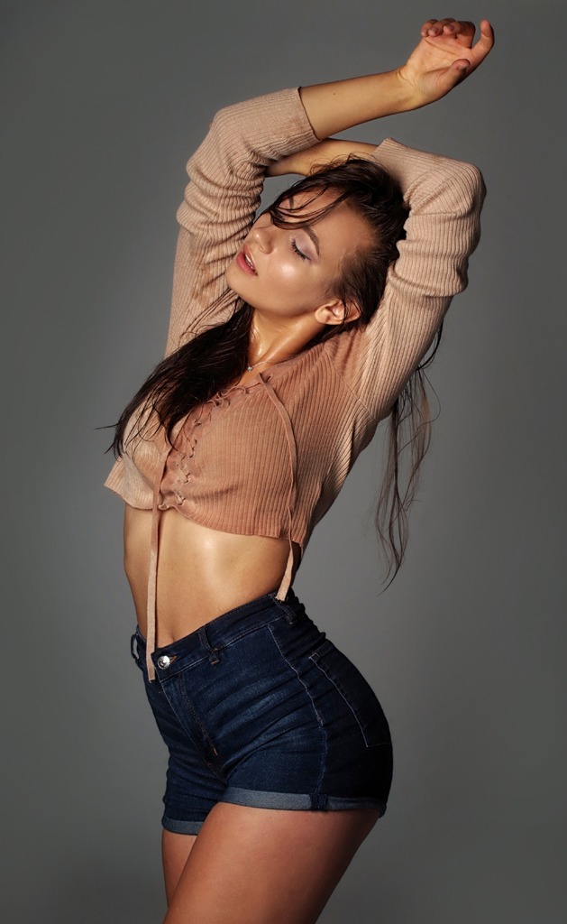 Mokra brunetka w krótkim topie i dżinsowych szortach, pozuje z uniesionymi rękami na szarym tle podczas sesji kobiecej.