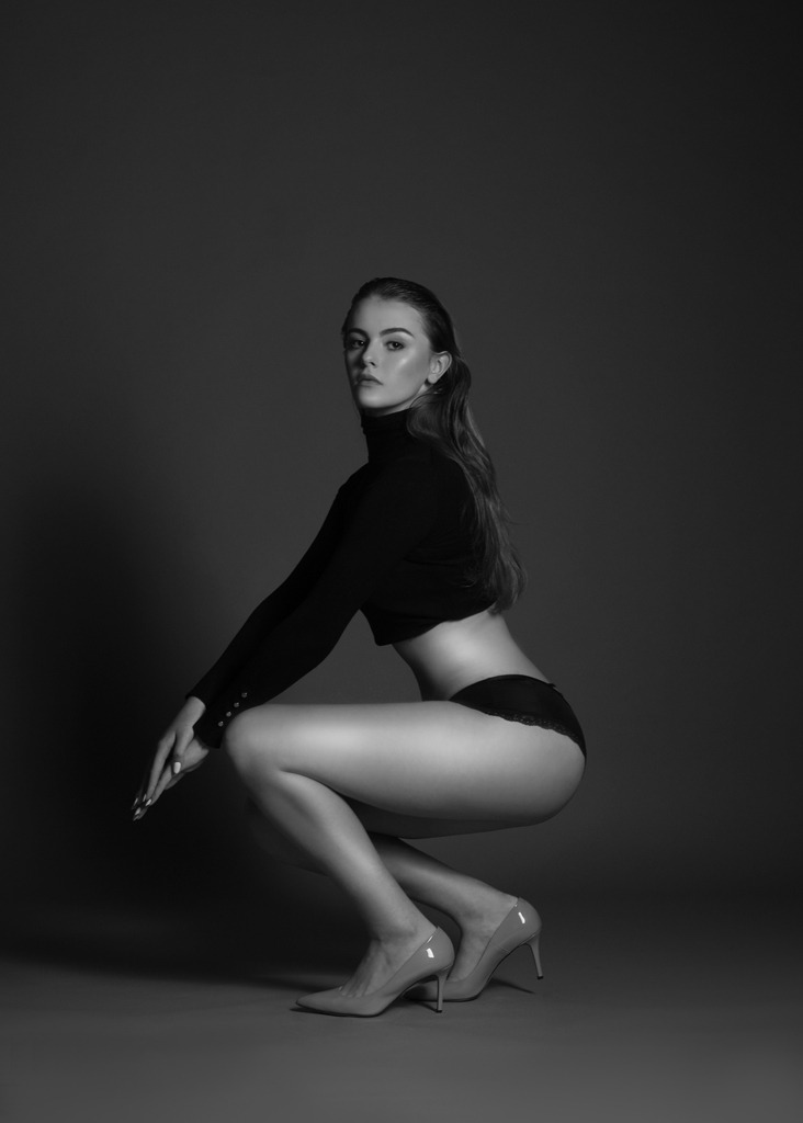 Czarno-białe zdjęcie w Łodzi przedstawia pozującą, szczupłą kobietę w czarnym golfie z długim rękawem, czarnych majtkach z koronką i beżowych szpilkach na wysokim obcasie, kucającą na szarym tle.