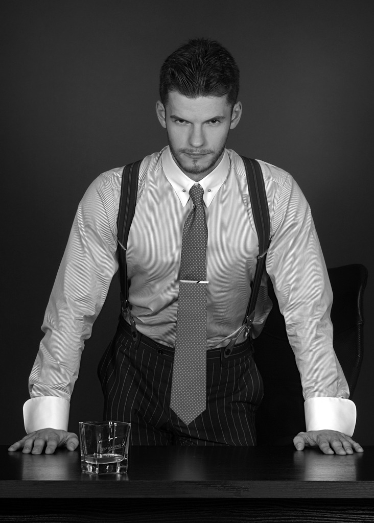 Mężczyzna w koszuli w paski, spodniach w prążki i szelkach, oparty o biurko z szklanką, w czarno-białej tonacji.