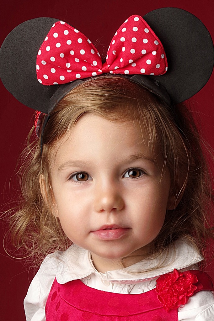 Portret uśmiechniętej dziewczynki z brązowymi oczami, w opasce z uszami Myszki Miki z czerwoną kokardą w białe kropki, na czerwonym tle.