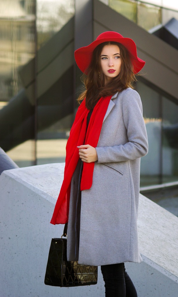 Portret młodej kobiety w szarym płaszczu, czerwonym kapeluszu i szalu, pozującej na tle nowoczesnej architektury, trzymającej czarną lakierowaną torebkę z geometrycznym wzorem.
