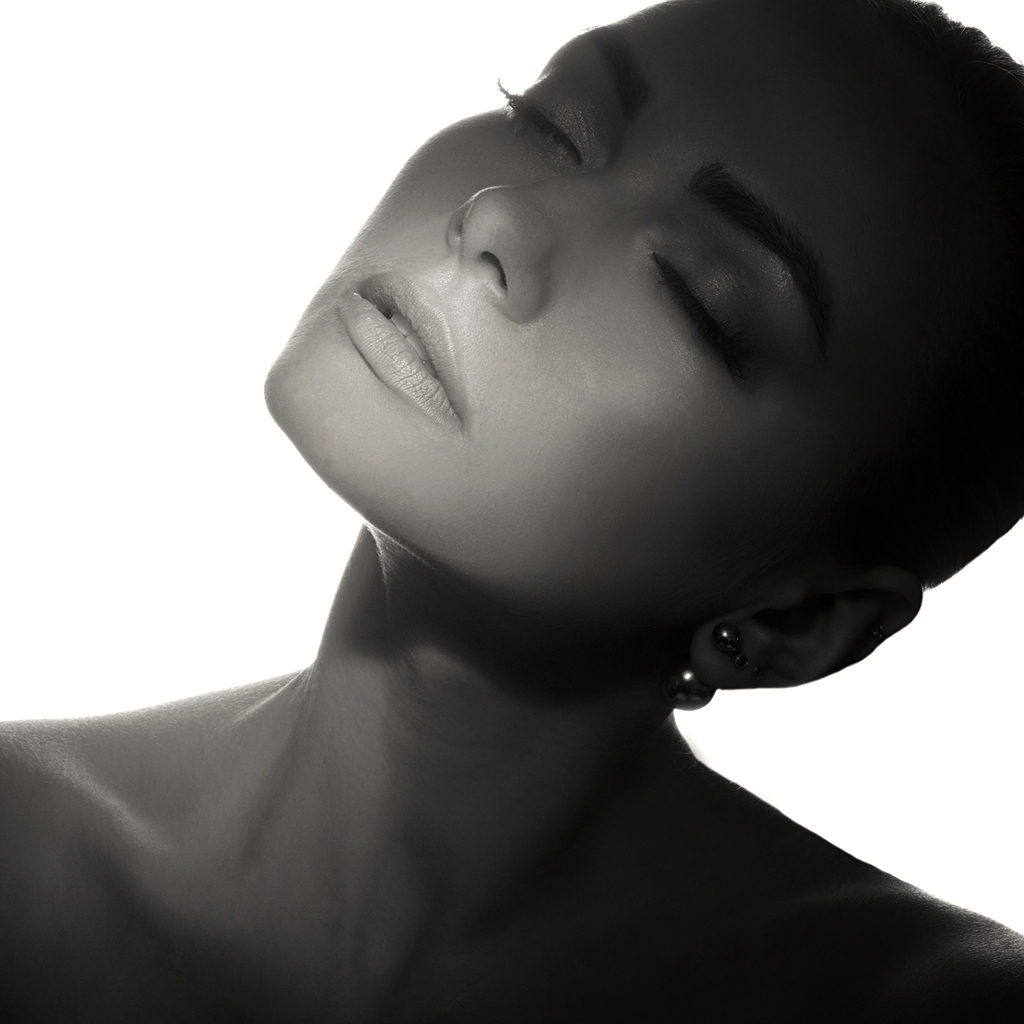 Czarno-biały portret kobiety z zamkniętymi oczami, skierowanej twarzą do góry, z delikatnym makijażem i kolczykami w uszach, oświetlony od tyłu.