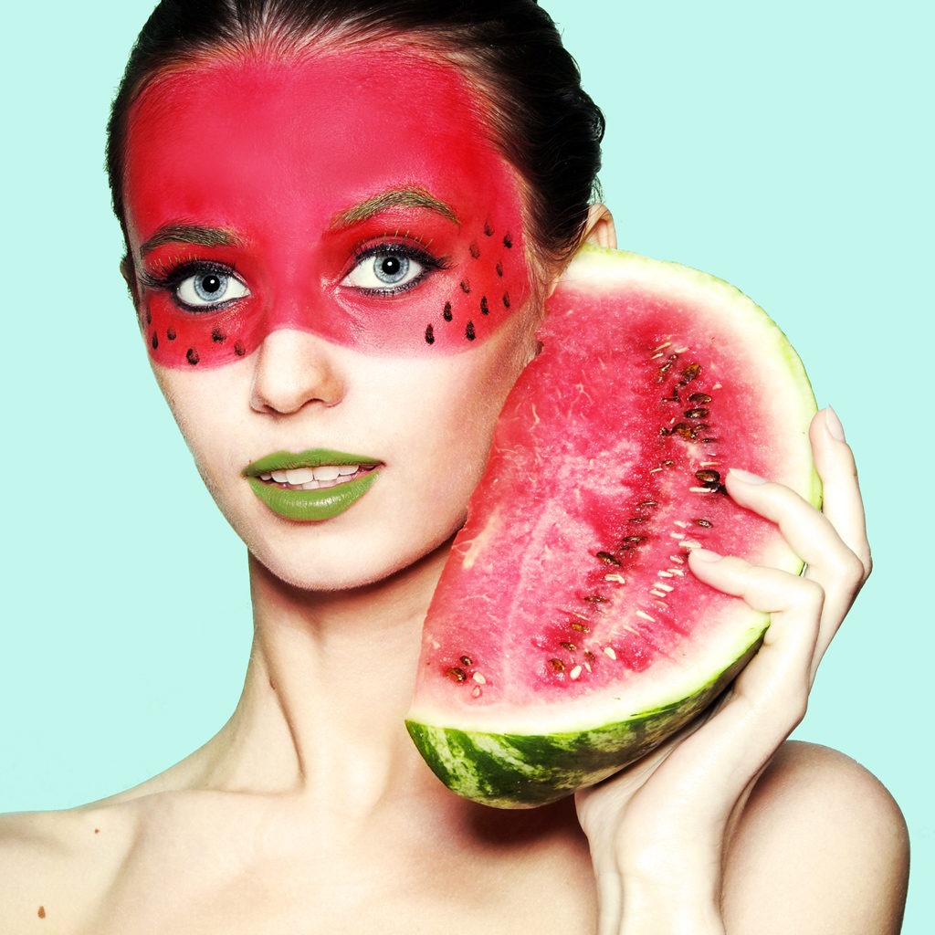 Portret kobiety z makijażem imitującym arbuza i trzymającej plaster arbuza przy twarzy, usta pomalowane na zielono, jasnoniebieskie tło.