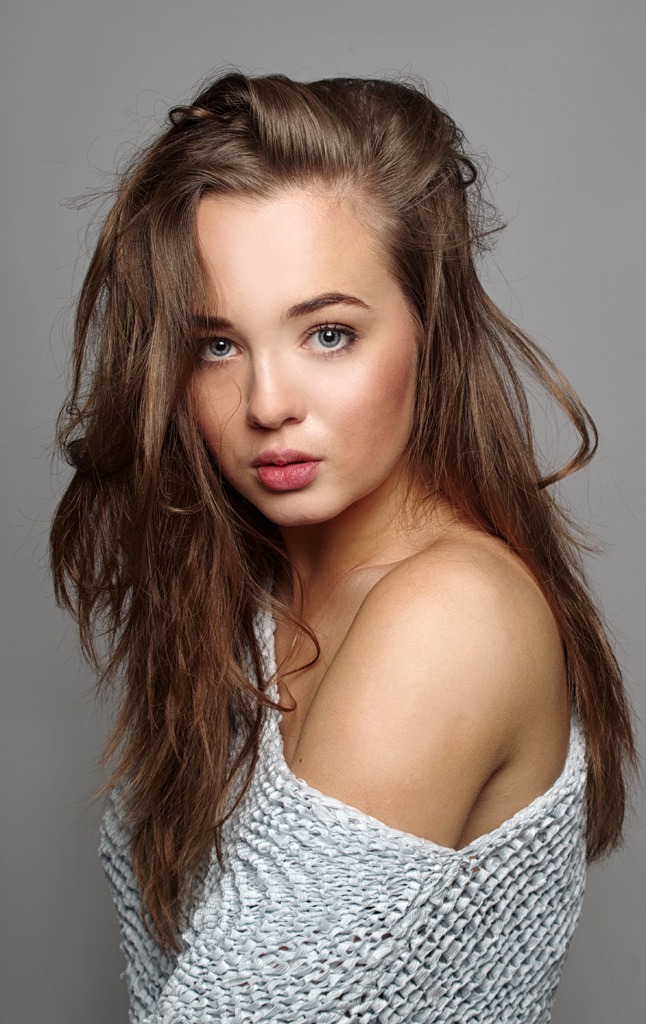 Portret młodej kobiety z rozwianymi włosami, w dzierganym topie na szarym tle.