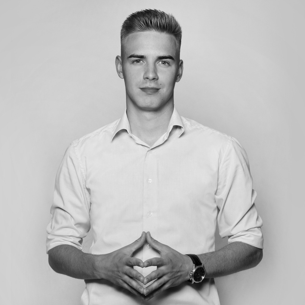 Portret młodego mężczyzny w białej koszuli na jednolitym tle, dłonie złożone przed sobą, widoczny zegarek na skórzanym pasku, zdjęcie czarno-białe.
