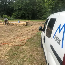 Wykonywanie fundamentów pod dom jednorodzinny, widoczny pracownik przy deskowaniu oraz samochód dostawczy z logo firmy na tle zieleni.