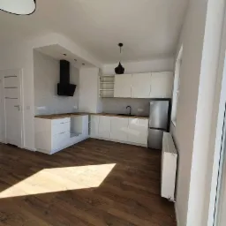 Nowoczesna, jasna kuchnia z białymi, lakierowanymi szafkami, drewnianym blatem, czarną armaturą i okapem, oraz drewnopodobną podłogą w nowym mieszkaniu, oświetlona wiszącą lampą.