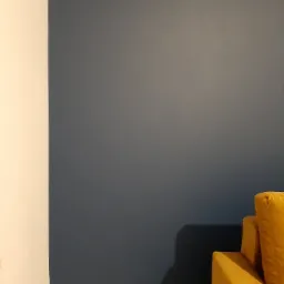 Fragment pokoju z granatową ścianą, białą listwą przypodłogową i narożnikiem sofy w kolorze musztardowym.