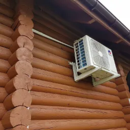 Biała jednostka zewnętrzna klimatyzatora marki Gree zamontowana na brązowej, drewnianej ścianie domu z bali, z widoczną anteną satelitarną w tle.