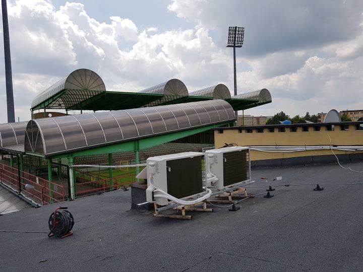 Dwa klimatyzatory zewnętrzne zamontowane na dachu stadionu GKS Bełchatów, widoczne zadaszenia trybun i maszt oświetleniowy w tle.