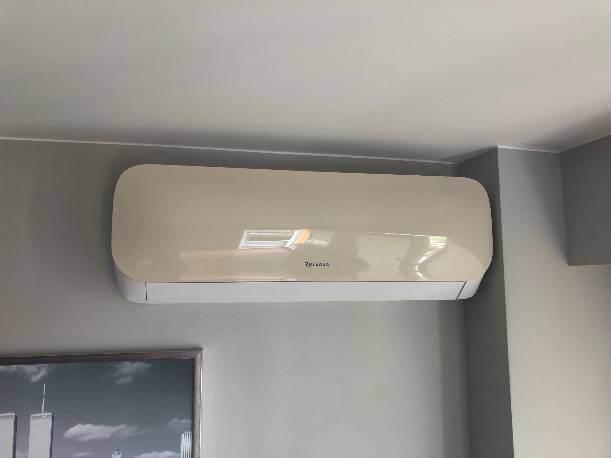 Zamontowany klimatyzator Rotenso na szarej ścianie, z widocznym odbiciem okna i fragmentem obrazu z wieżami World Trade Center w dolnym rogu.