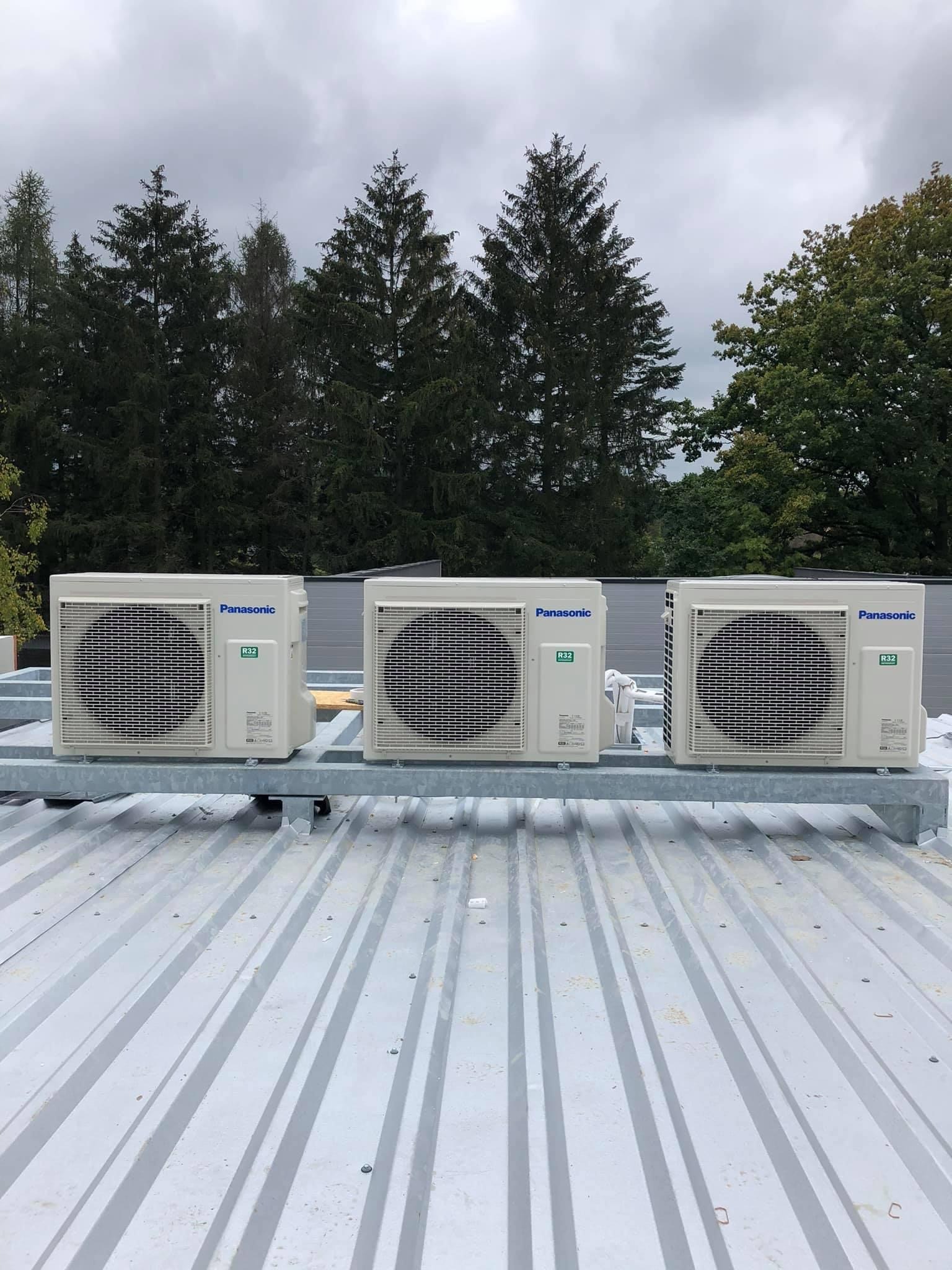 Trzy klimatyzatory marki Panasonic zamontowane na metalowej konstrukcji na dachu, widok z dołu na tle drzew i pochmurnego nieba.