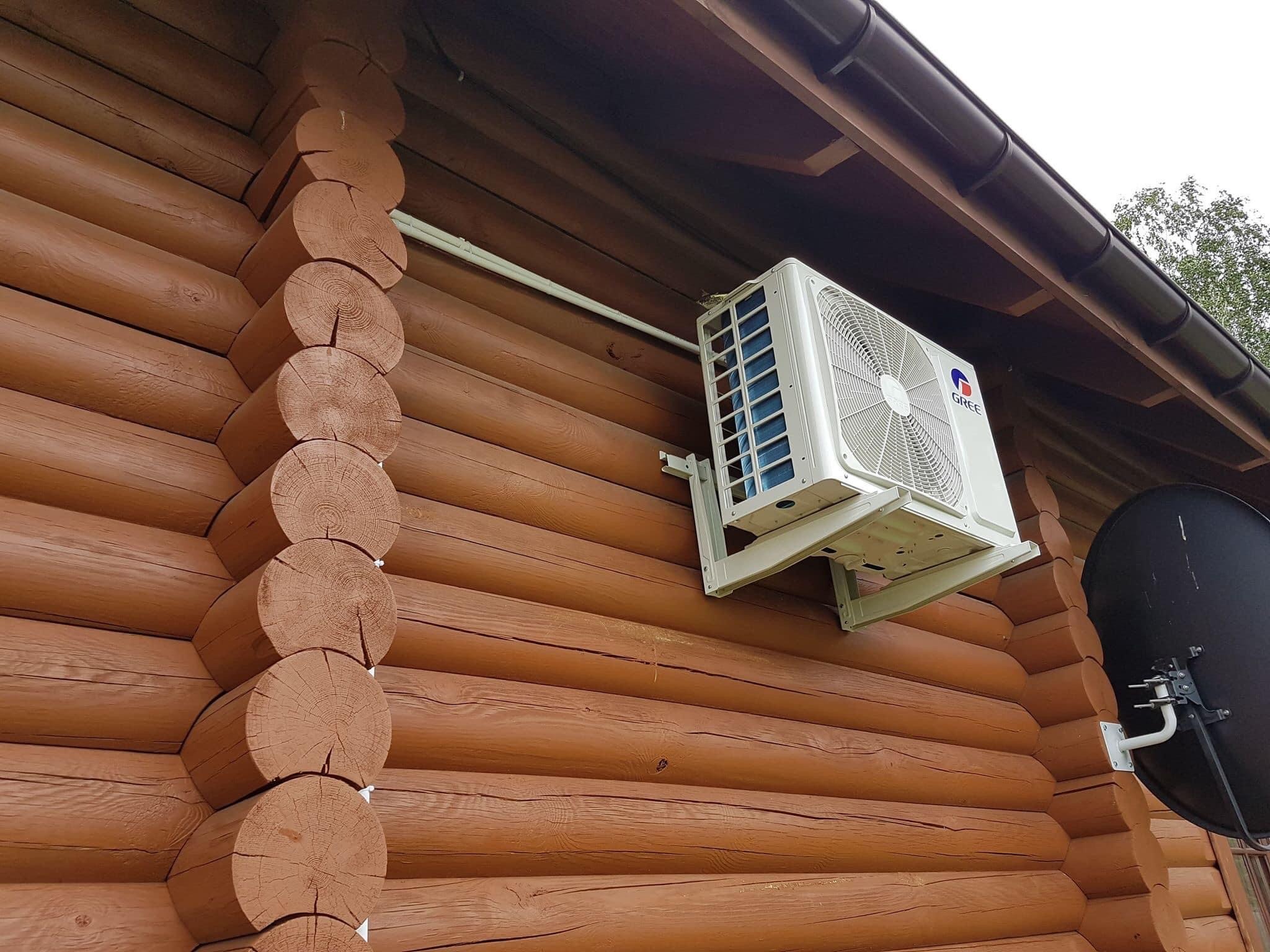 Biała jednostka zewnętrzna klimatyzatora marki Gree zamontowana na brązowej, drewnianej ścianie domu z bali, z widoczną anteną satelitarną w tle.