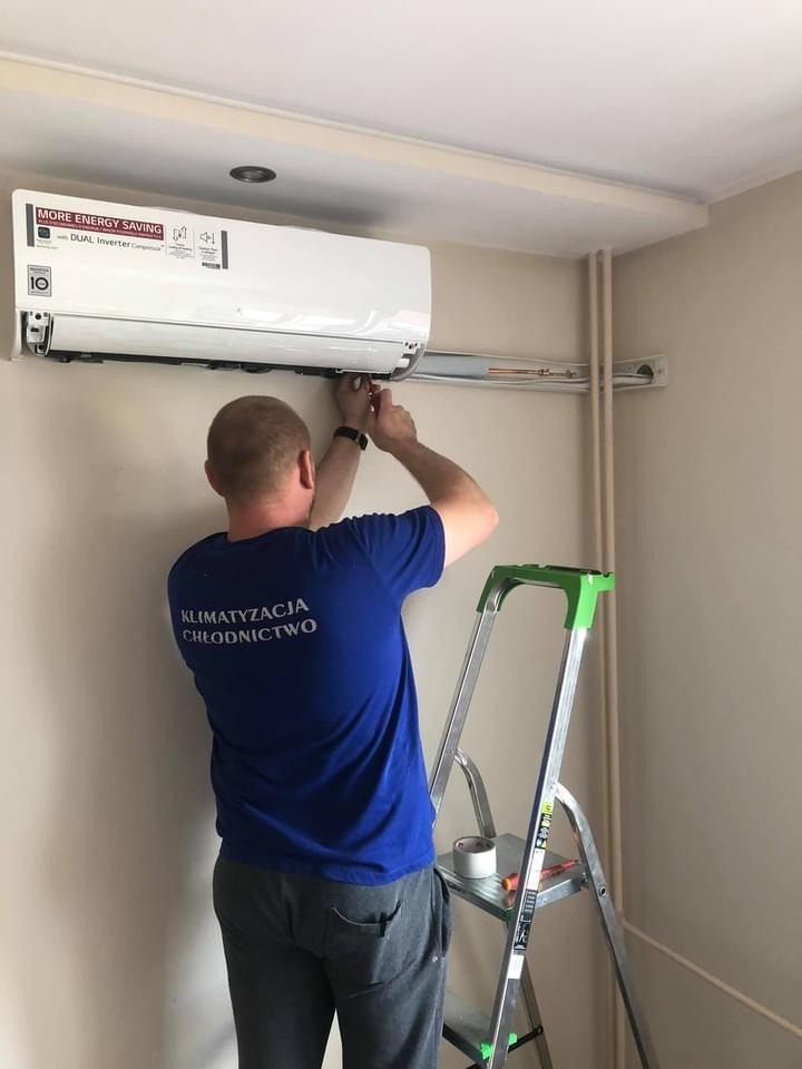 Instalator w niebieskiej koszulce firmowej montuje klimatyzator na ścianie w jasnym pomieszczeniu, stojąc na aluminiowej drabinie z zielonymi elementami.