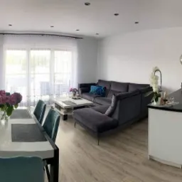 Panoramiczne ujęcie nowocześnie urządzonego wnętrza z otwartą kuchnią, jadalnią i salonem, charakteryzujące się jasną kolorystyką i drewnianą podłogą, z akcentami turkusowych krzeseł i szarej sofy.