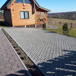 Przepuszczalna nawierzchnia parkingowa z ażurowych płyt betonowych wypełnionych drobnym żwirem, przylegająca do drewnianego domu z balkonem i ścieżki z kostki, w tle krajobraz podmiejski.