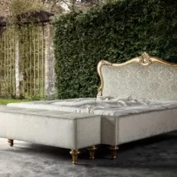 Tapicerowane łóżko z wezgłowiem w stylu glamour, ozdobione złotymi detalami, ustawione na zewnątrz na tle zielonej ściany z roślin.