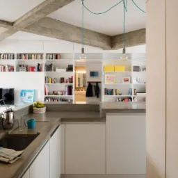 Nowoczesna kuchnia z betonowym blatem, białymi szafkami i półkami z książkami w tle, oświetlona wiszącymi lampami na zielonych kablach.