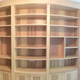 Drewniana zabudowa ściany z półkami i szafkami, z widocznym blatem biurka przy oknie, wykonana z jasnego drewna o wyraźnym usłojeniu.