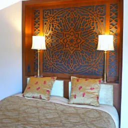 Łóżko z ozdobnym, ażurowym wezgłowiem z drewna w stylu orientalnym, oświetlone dwiema lampkami nocnymi z abażurami, na łóżku leżą dwie poduszki z motywem kwiatowym i beżowa narzuta.