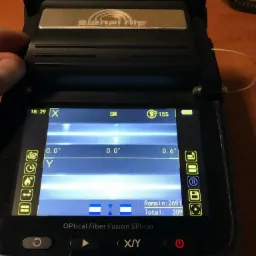 Otwarty splicer światłowodowy z widocznym ekranem LCD prezentującym parametry spawania, w tle widoczny fragment pulpitu.