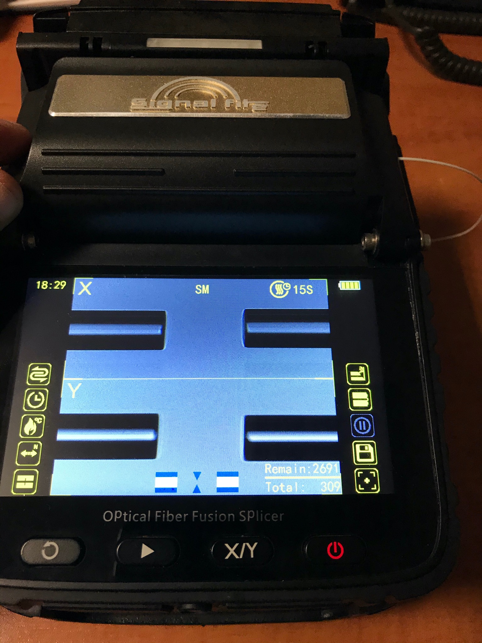 Otwarty spawacz światłowodowy Signal Fire z widocznym ekranem wyświetlającym proces łączenia włókien. Na wyświetlaczu widoczne parametry spawania oraz ikony funkcyjne. W tle widoczny kabel...