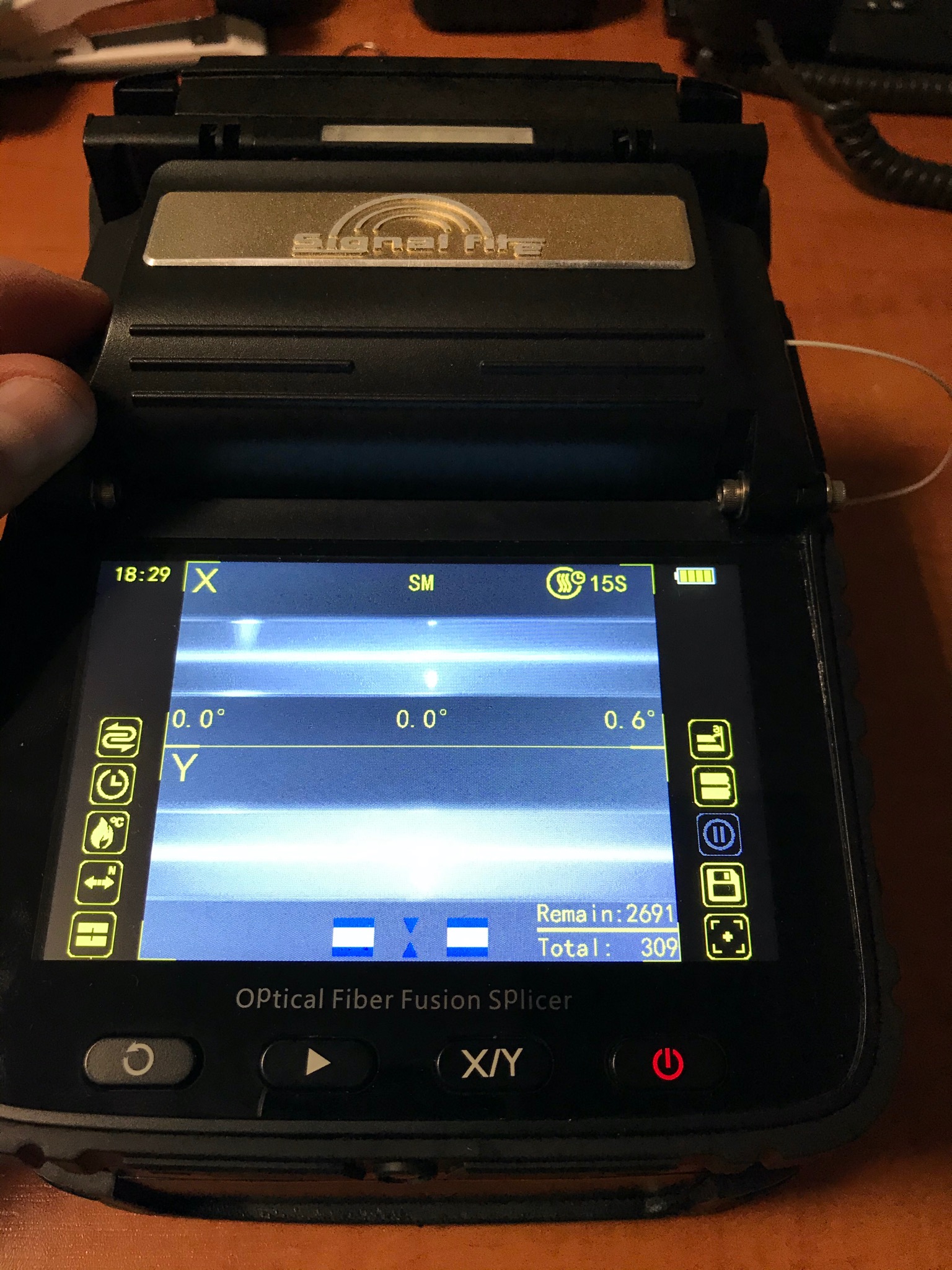 Otwarty splicer światłowodowy z widocznym ekranem LCD prezentującym parametry spawania, w tle widoczny fragment pulpitu.